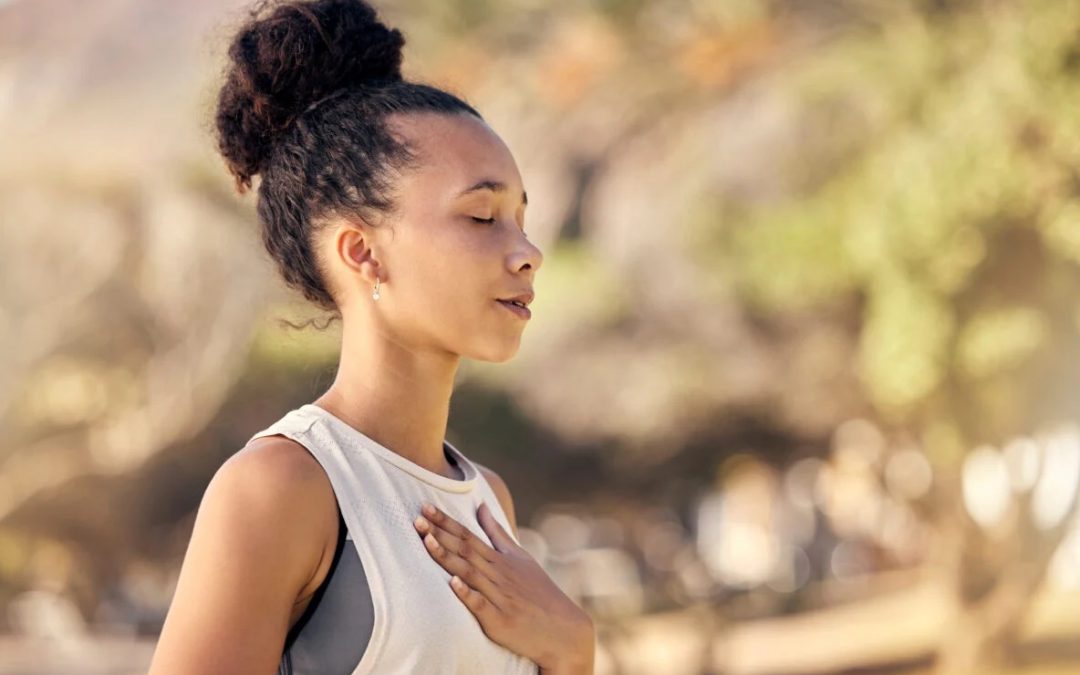 Woman practicing breathing exercises outdoors, with one hand on her chest and her eyes closed, appearing focused and restored.